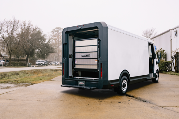 White delivery van with an open rear liftgate, parked on a concrete area, surrounded by trees and a building in the background.